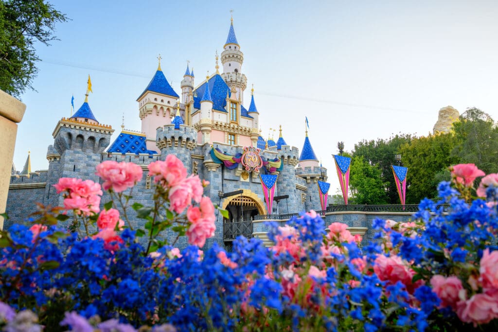 The Disneyland Resort Sleeping Beauty Castle is adorned with 70th Celebration decor and pink, blue and purple flowers in the foreground.