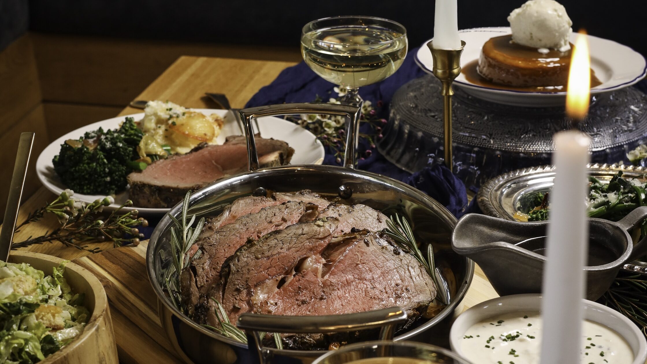 A prime rib meal with plates of broccoli, bowls of salad, sauces and lit candle on the table at Doughbird in Arizona.