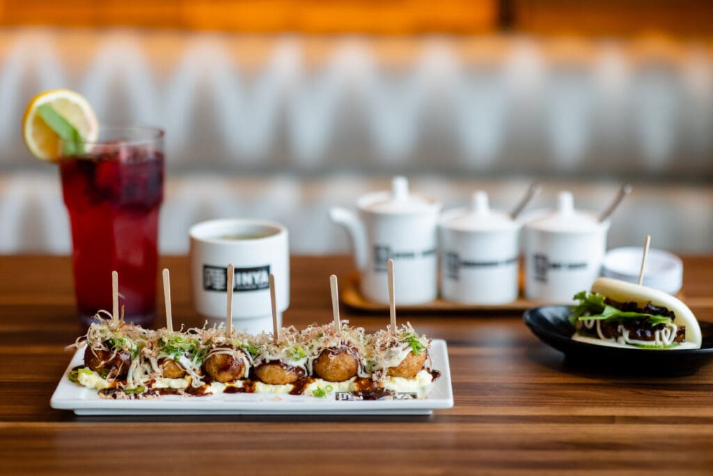 A row of appetizers on a rectangular plate and other app with a garnished drink and JINYA branded tableware in the background at JINYA Ramen Bar.