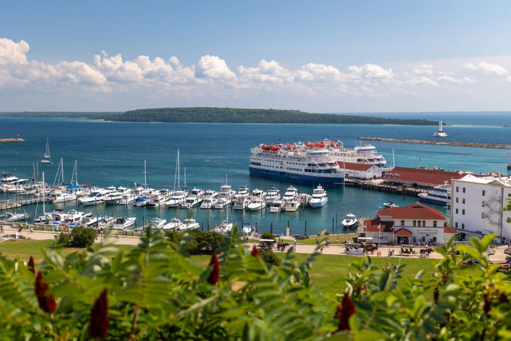 A view of the marina with sailboats, ferries, people walking and riding horse-drawn carriages on a marina road on Mackinac Island Tourism Bureau.