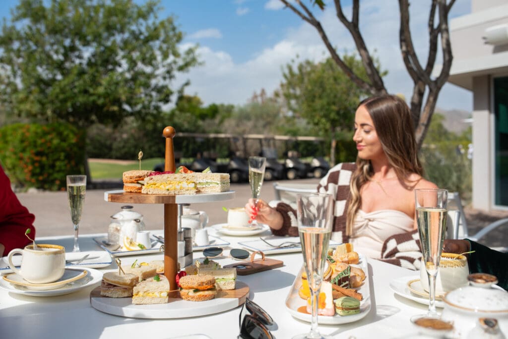 A woman holds a Champagne flute with tea party food set on trays at Mountain Shadows Resort.