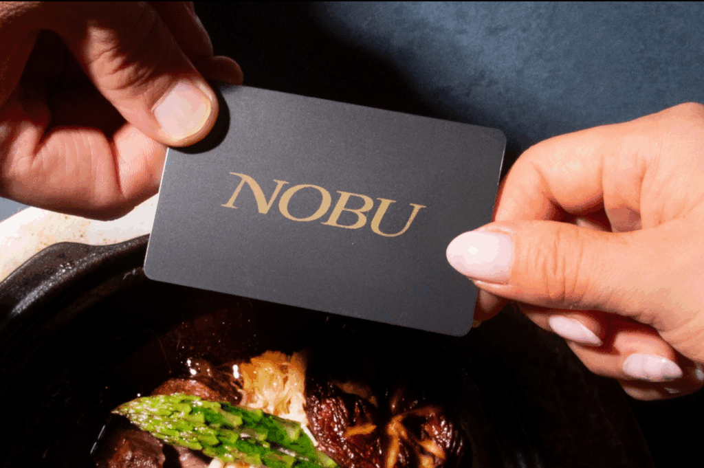 Two hands hold a Nobu gift card with a Japanese food dish underneath.