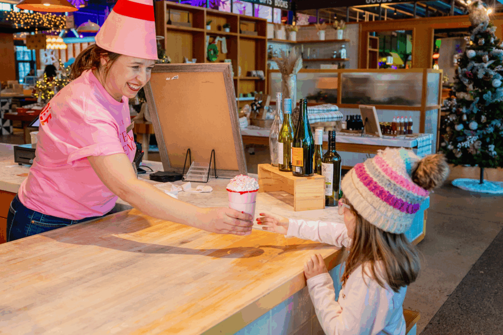 A woman in a pink elf outfit hands a pink-whip drink to a little girl wearing glasses and a beanie during the holiday season at Sawmill Market.