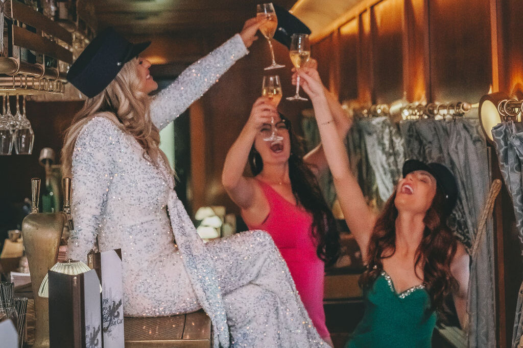Three women in NYE cocktail attire and conductor hats hold up three glasses of champagne at Grey Hen, Century Grand in Phoenix. 