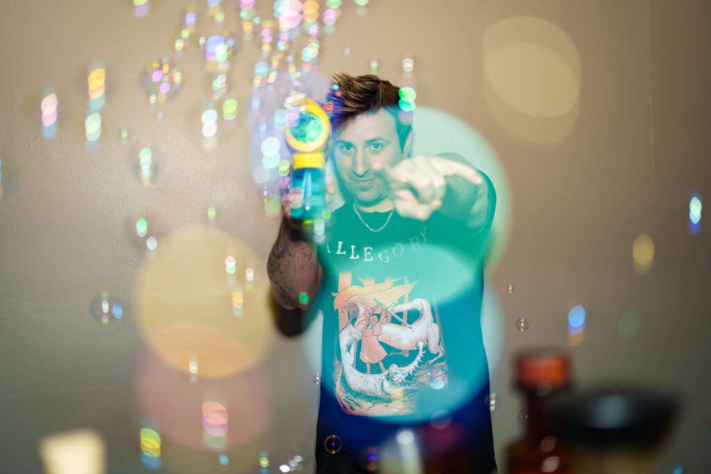 A man points to the camera, holding a handheld bubble machine expelling bubbles at Arizona Cocktail Weekend. 