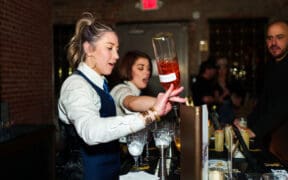 A bartender pours a bottle of liquor into glass with another bartender in the background making a drink at Arizona Cocktail Weekend.