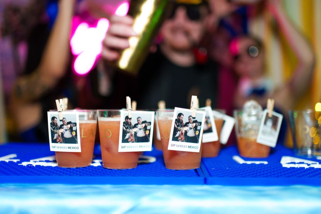 A row of sample plastic cups with cocktails have photos of two men attached to them with clothes pins at Arizona Cocktail Weekend.